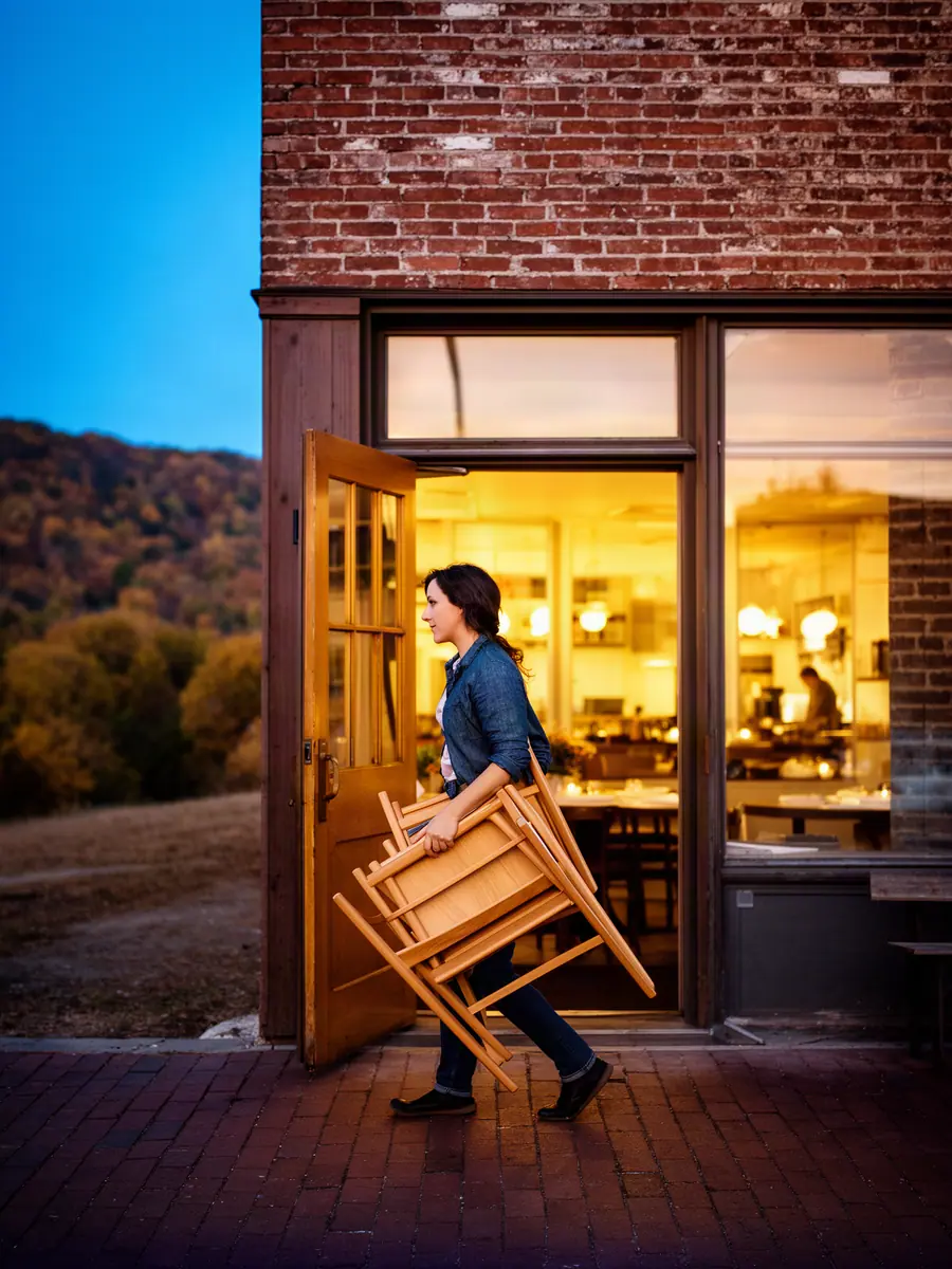A restaurant owner carrying folding chairs out of her warm-lit front door onto a brick sidewalk at dusk.